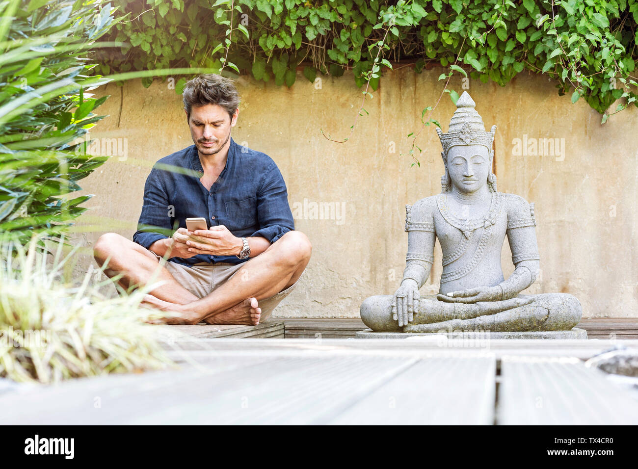 Homme assis en tailleur à côté de la statue de Bouddha dans un jardin Zen, using smartphone Banque D'Images