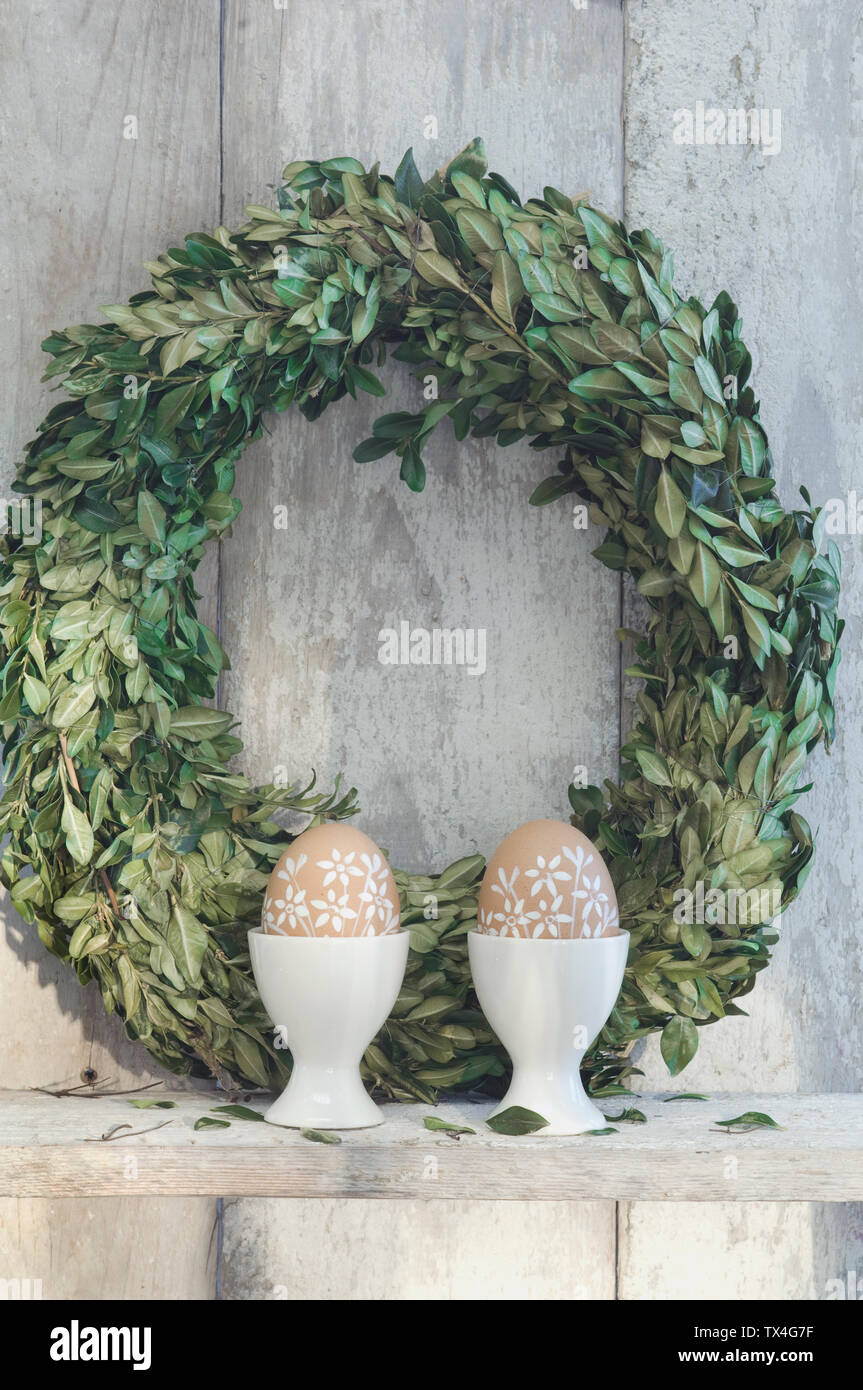 Deux oeufs de Pâques peints dans les tasses et couronne de buis sur la tablette en face de mur en bois Banque D'Images