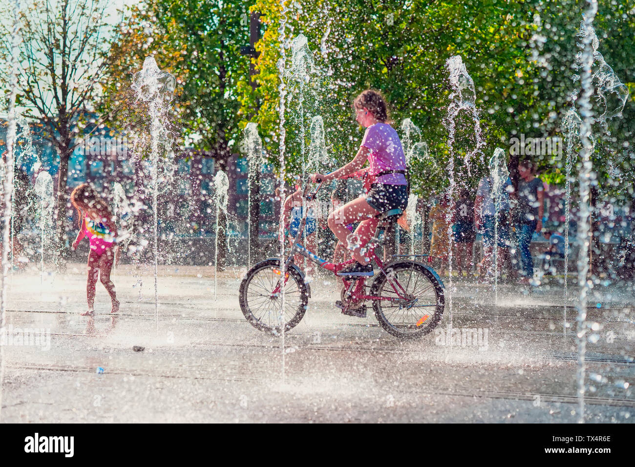 L'été, la chaleur, une fille faire du vélo entre les fontaines, les gens cool dans des jets d'eau Banque D'Images