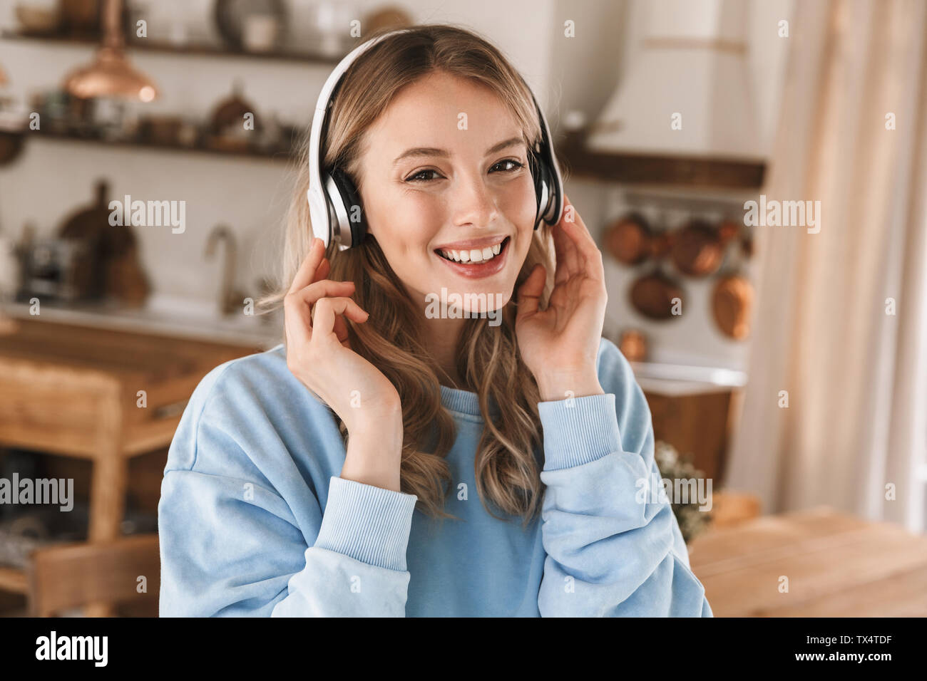 Portrait de jeune fille blonde moderne 20s portant des écouteurs de sourire et de joie tout en écoutant de la musique à la maison Banque D'Images