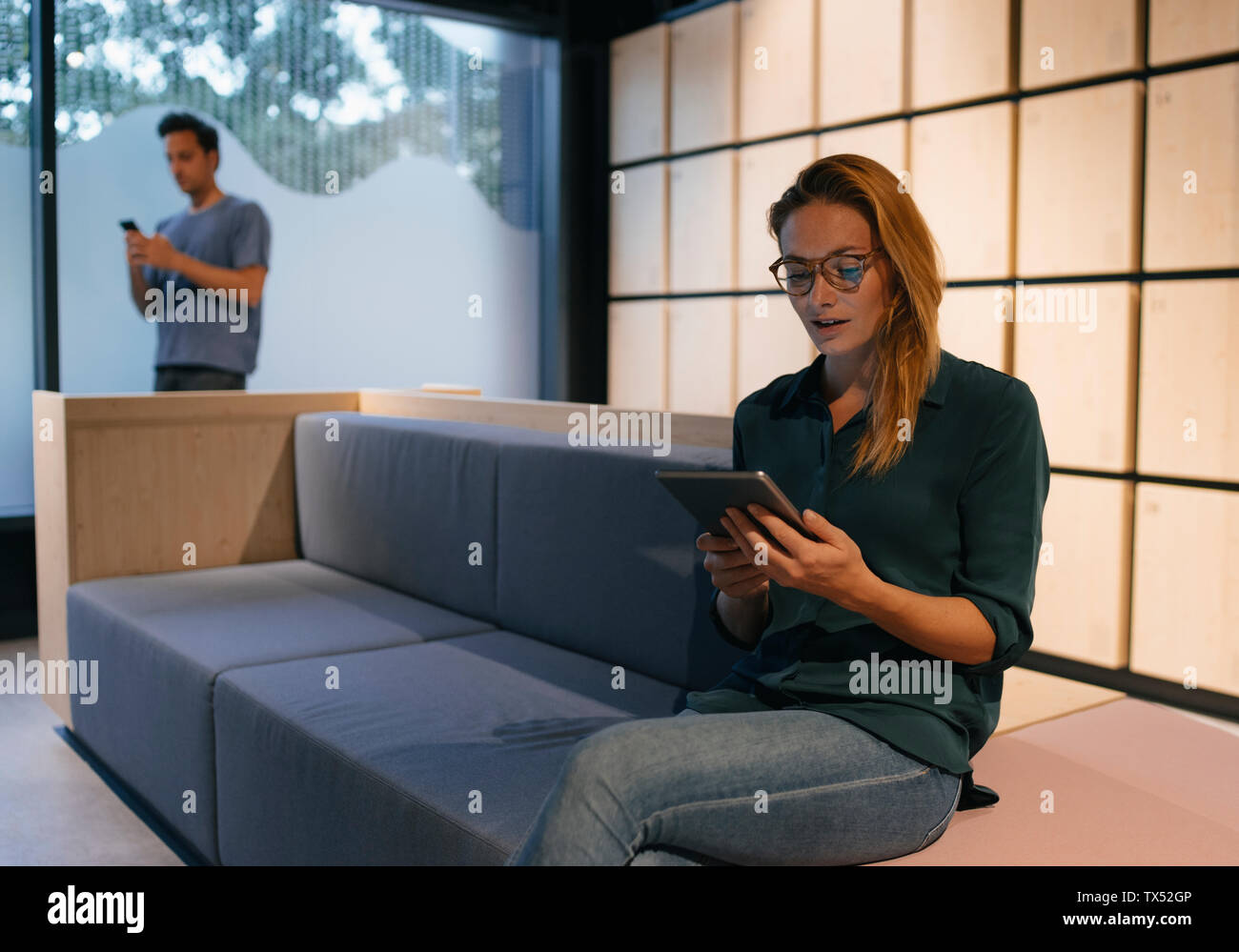 Young woman sitting on couch using tablet avec l'homme en arrière-plan Banque D'Images