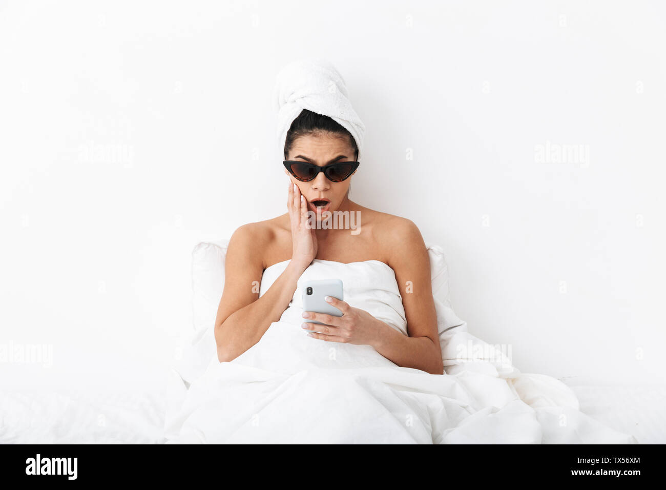 Image d'une belle surprise avec femme émotionnelle towel on head se trouve au lit sous couverture isolated over white wall background portant des lunettes de cha Banque D'Images