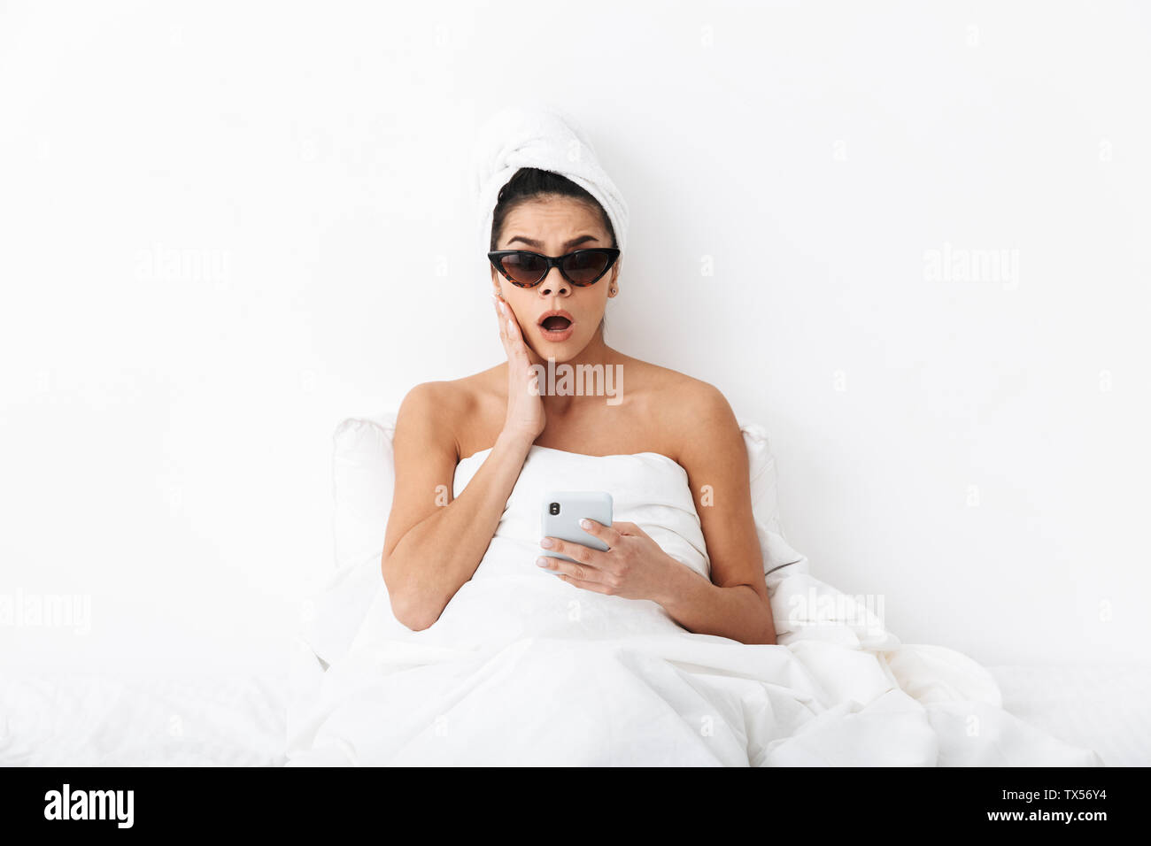 Image d'une belle surprise avec femme émotionnelle towel on head se trouve au lit sous couverture isolated over white wall background portant des lunettes de cha Banque D'Images