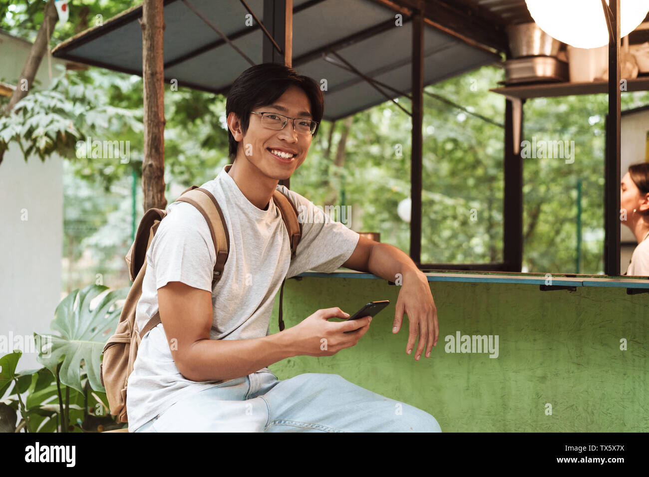 Smiling asian student homme portant un sac à dos à l'extérieur, assis, à l'aide de mobile phone Banque D'Images