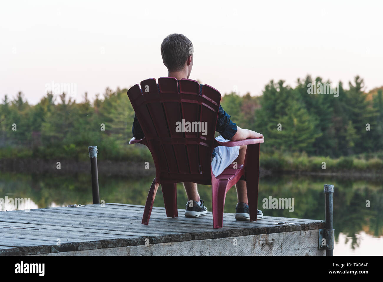 Jeune homme de vous détendre sur une chaise Adirondack et à la recherche à une rivière calme au coucher du soleil. Muskoka, Ontario, Canada. Banque D'Images