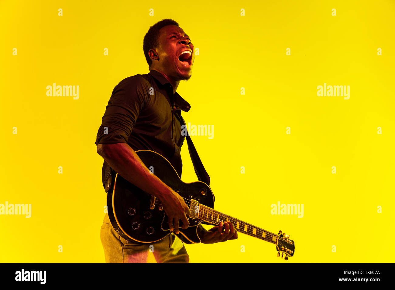 Les jeunes africains-américains musicien jouant de la guitare comme une rockstar sur fond jaune dans la région de neon light. Concept de la musique, passe-temps, festival, plein air. Guy attrayant joyeux improvisant, chantant la chanson. Banque D'Images