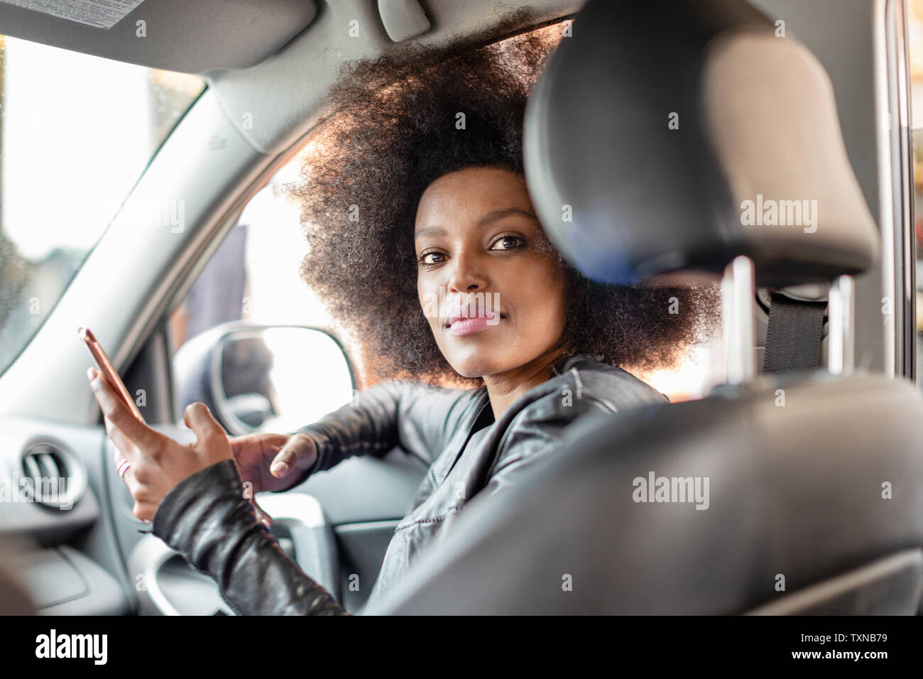 Jeune femme avec des cheveux afro en siège passager voiture holding smartphone, portrait Banque D'Images