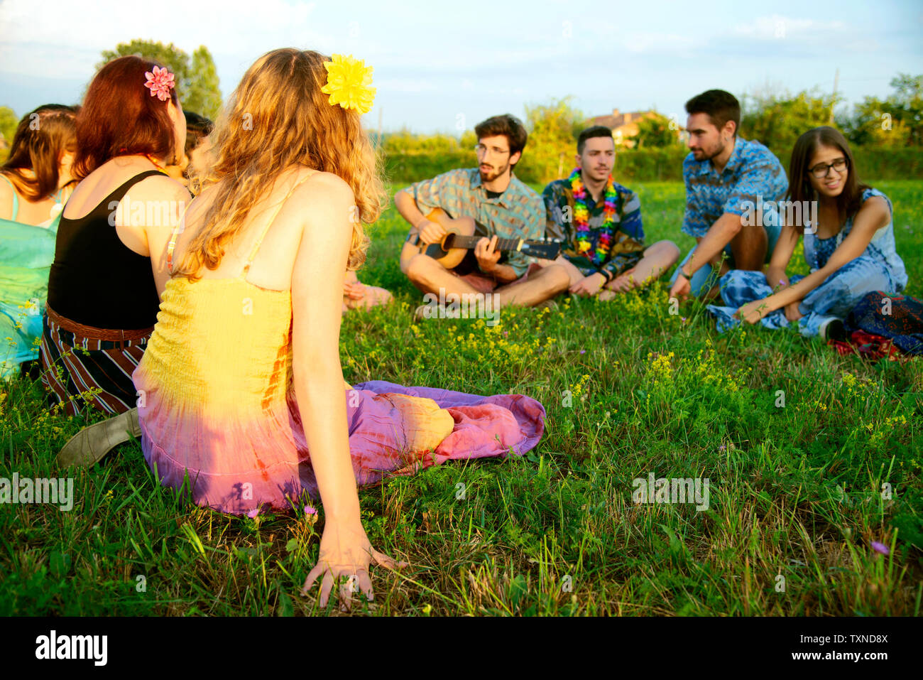 Grand groupe de jeunes adultes assis dans le champ à l'écoute de la guitare acoustique Banque D'Images