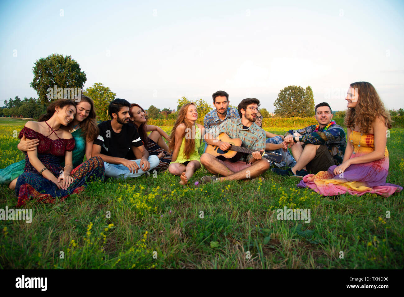 Grand groupe de jeunes adultes assis dans le champ à l'écoute de la guitare acoustique Banque D'Images