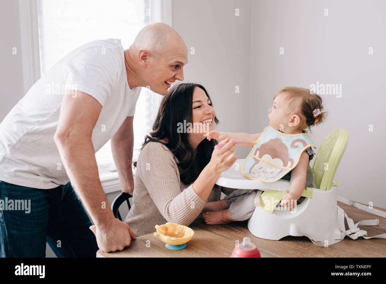 Mère et père nourrir bébé fille en siège enfant sur table de cuisine Banque D'Images