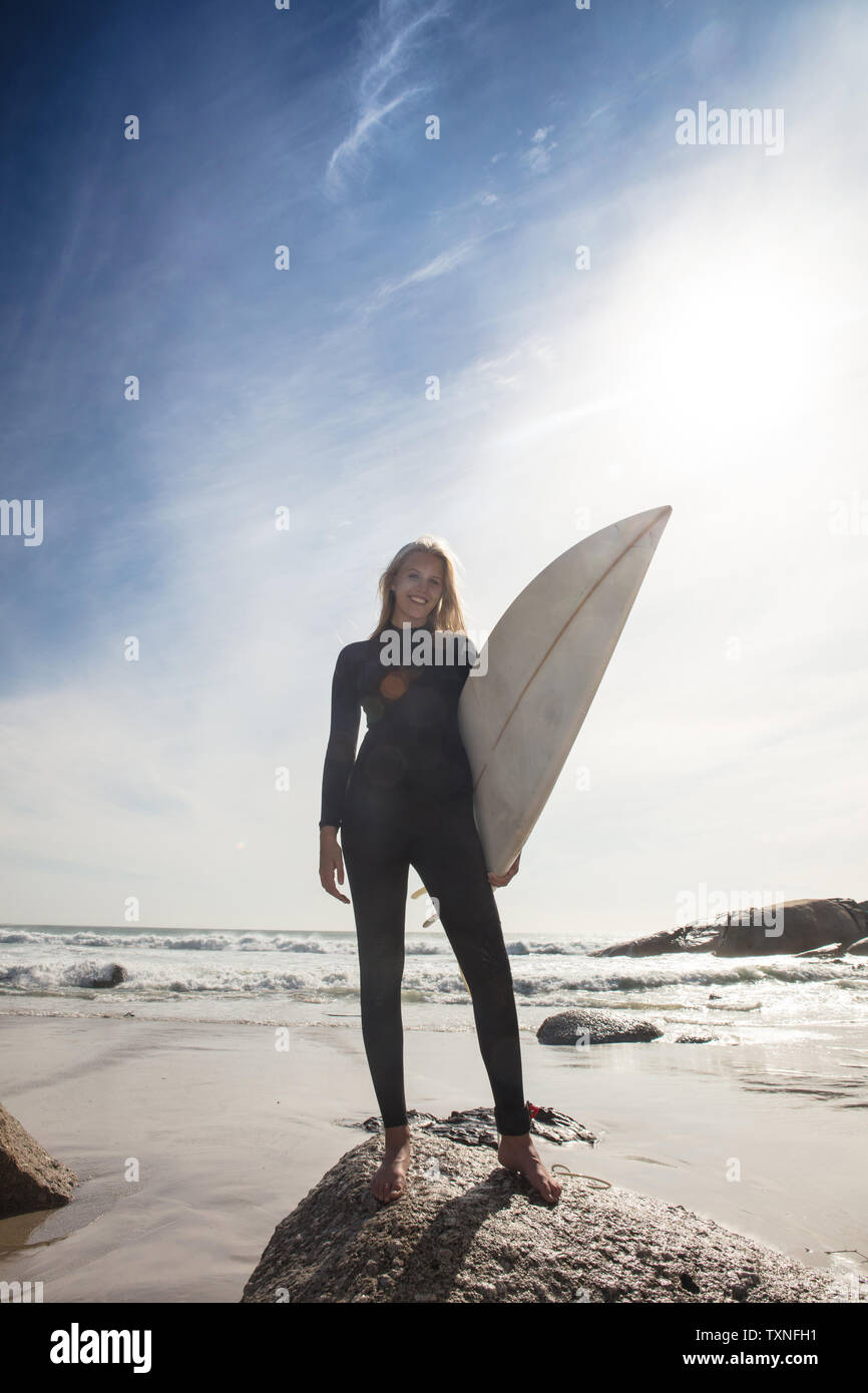 Jeune femme surfer carrying surfboard on beach rock, pleine longueur portrait, Cape Town, Western Cape, Afrique du Sud Banque D'Images