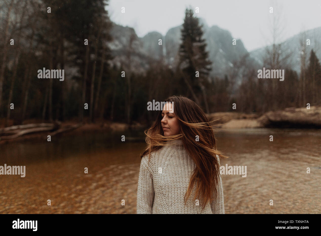 Jeune femme on riverbank shaking longs cheveux bruns, Yosemite Village, California, USA Banque D'Images
