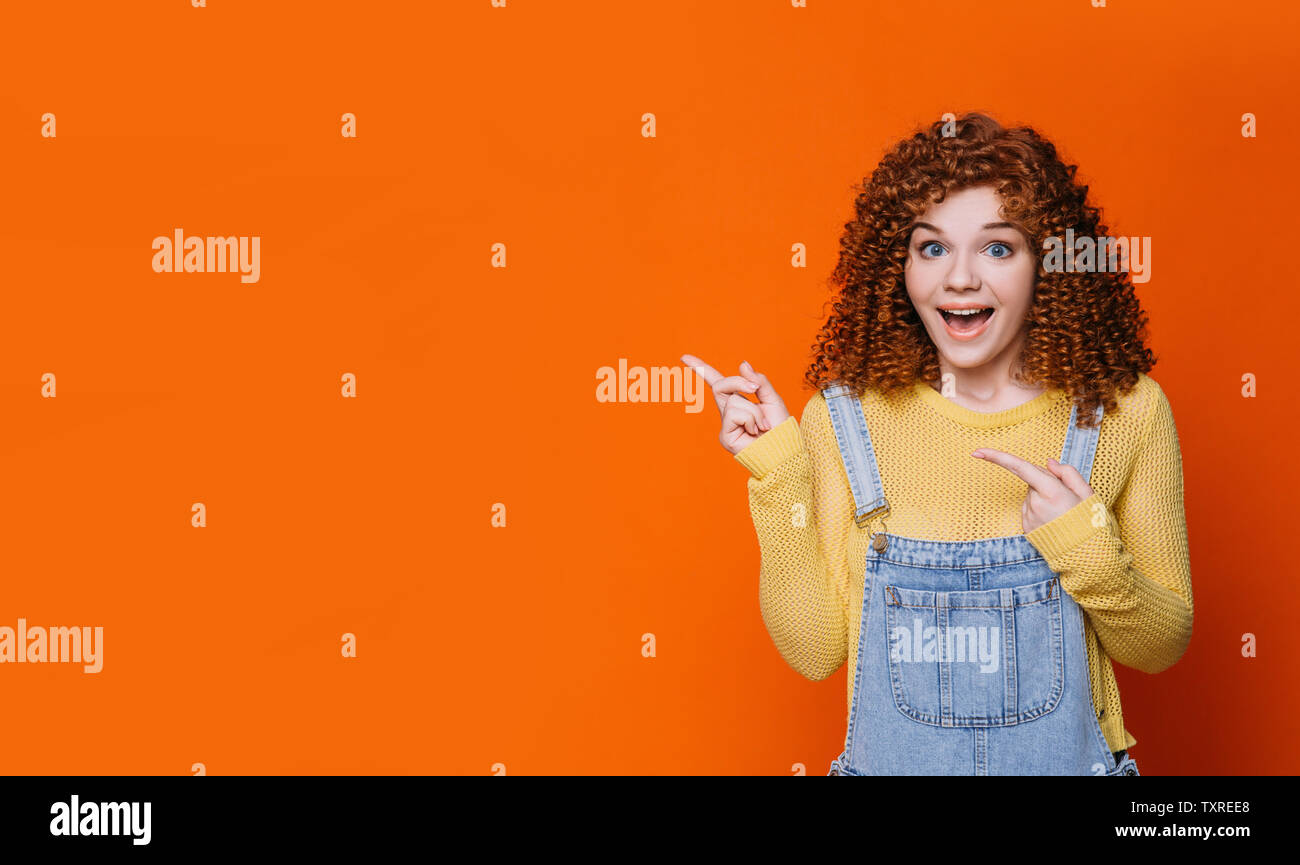 Curly red-haired woman faisant face surpris et en pointant les mains vides en fond orange. Wow incroyable Banque D'Images