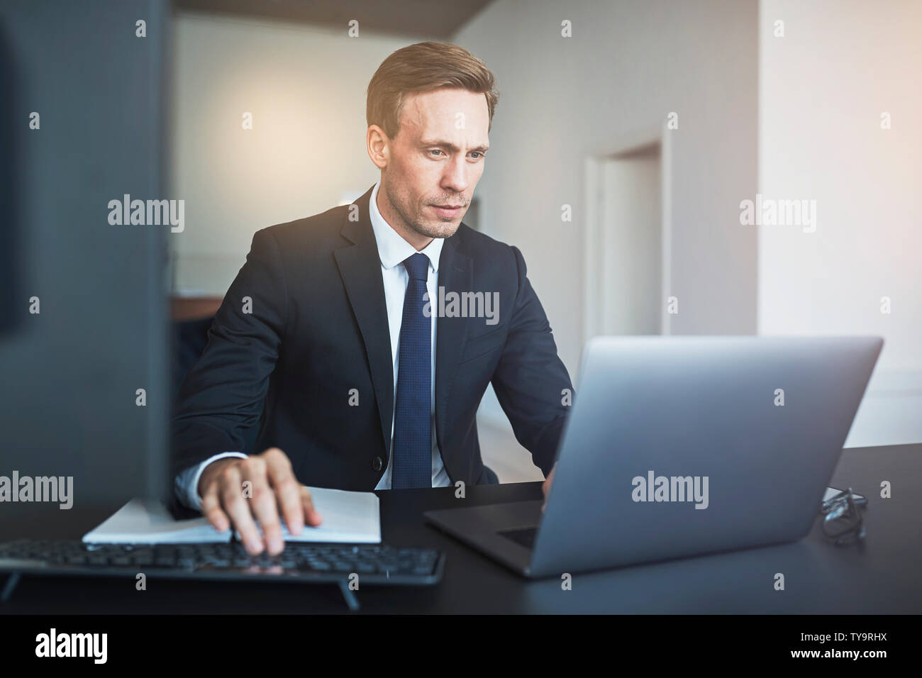 L'accent vertical en ligne avec un ordinateur portable tout en étant assis à son bureau dans un bureau moderne et lumineux Banque D'Images