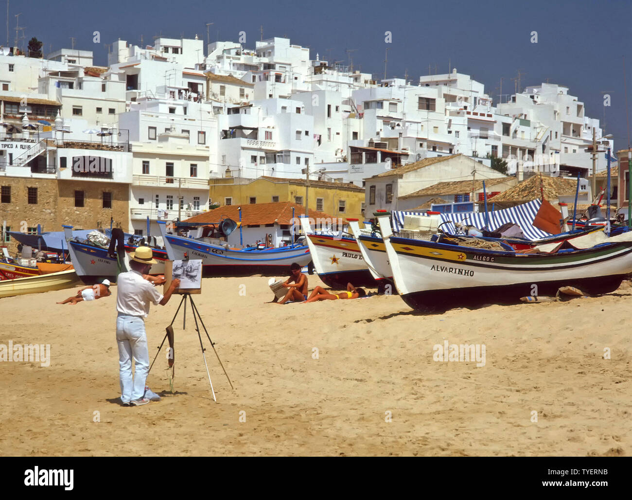 Man1980s artiste au travail sous ciel bleu clair avec tableau blanc esquisse sur maison de vacances soleil et plage de sable propre bateau de pêche Albufeira Algarve Portugal Banque D'Images