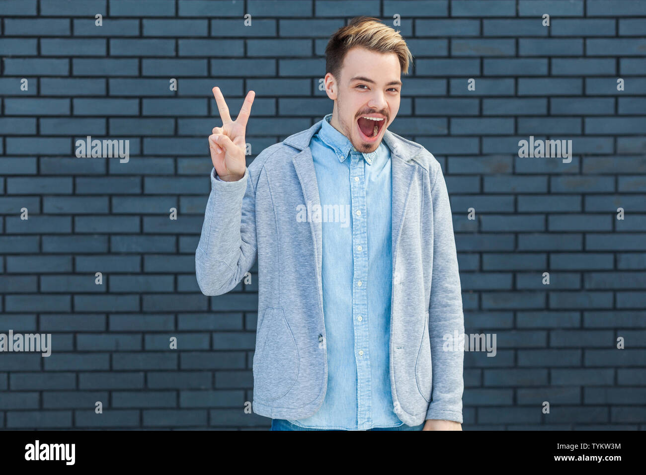 Portrait de surpris beau jeune homme blond au style casual debout avec la victoire ou la paix et geste looking at camera avec étonnement heureux face. je Banque D'Images