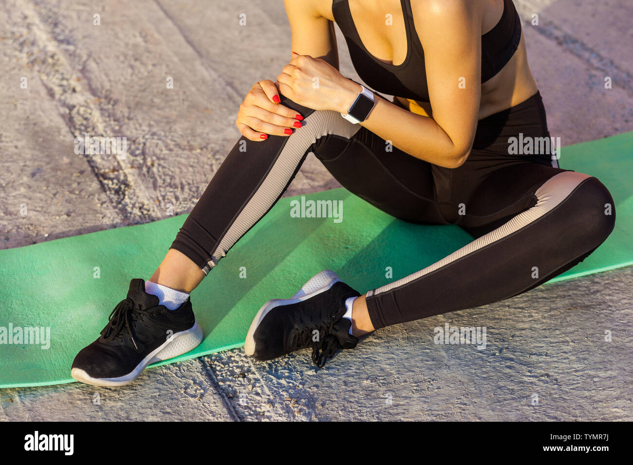 Closeup portrait of young woman in black sportwear assis sur tapis avec un problème au genou, mal avec spasme douloureux. tenant la jambe de préhension wit Banque D'Images