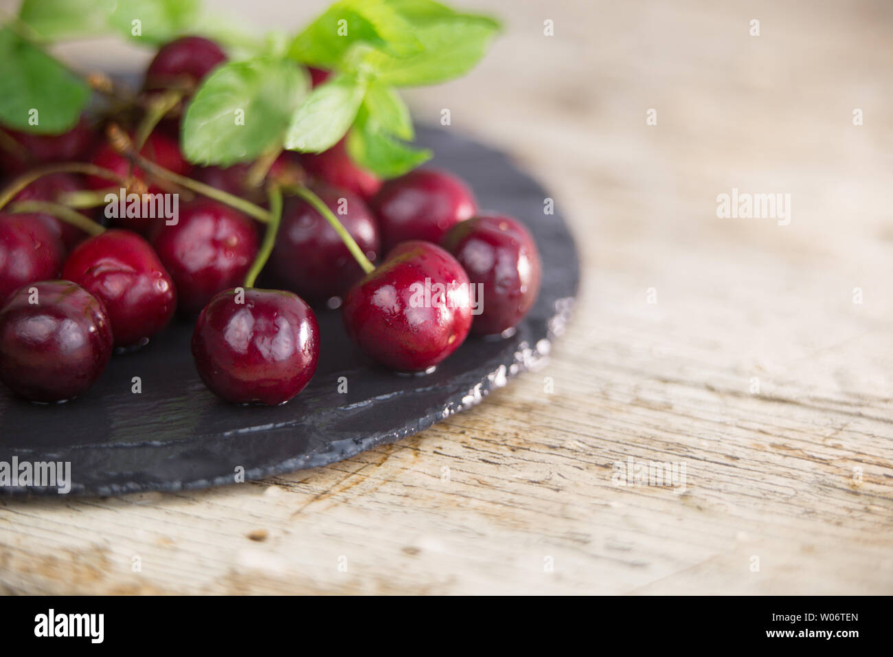 Cerises fraîches sur une plaque d'ardoise noire humide avec brin de menthe verte sur un parquet en bois clair table dans focus sélectif pour copy space Banque D'Images