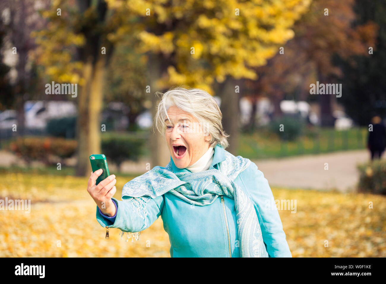 Vue d'un souriant et assez espressive senior woman looking at son téléphone sur feuilles jaunes en face d'un des arbres avec des couleurs automnales. Banque D'Images