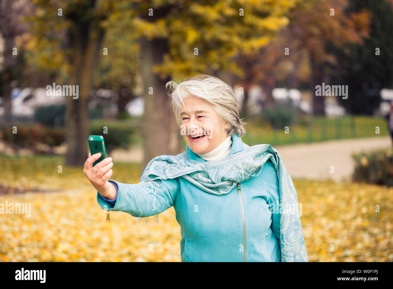 Vue d'un souriant et assez espressive senior woman looking at son téléphone sur feuilles jaunes en face d'un des arbres avec des couleurs automnales. Banque D'Images