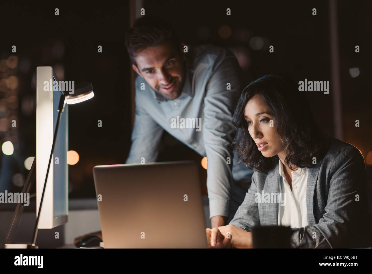 Deux jeunes gens d'affaires à l'aide d'un ordinateur portable dans un bureau 24 heures supplémentaires tout en travaillant ensemble en fin de soirée Banque D'Images