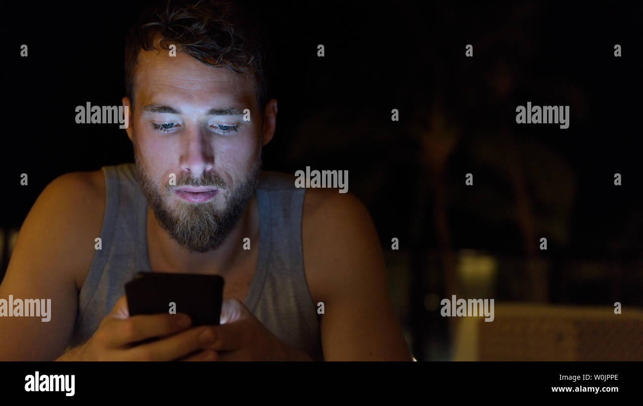 Man using smartphone à internet navigation de nuit mise à jour des médias sociaux. Jeune homme avec barbe à l'aide de mobile cell smart phone dans le noir en été. Banque D'Images