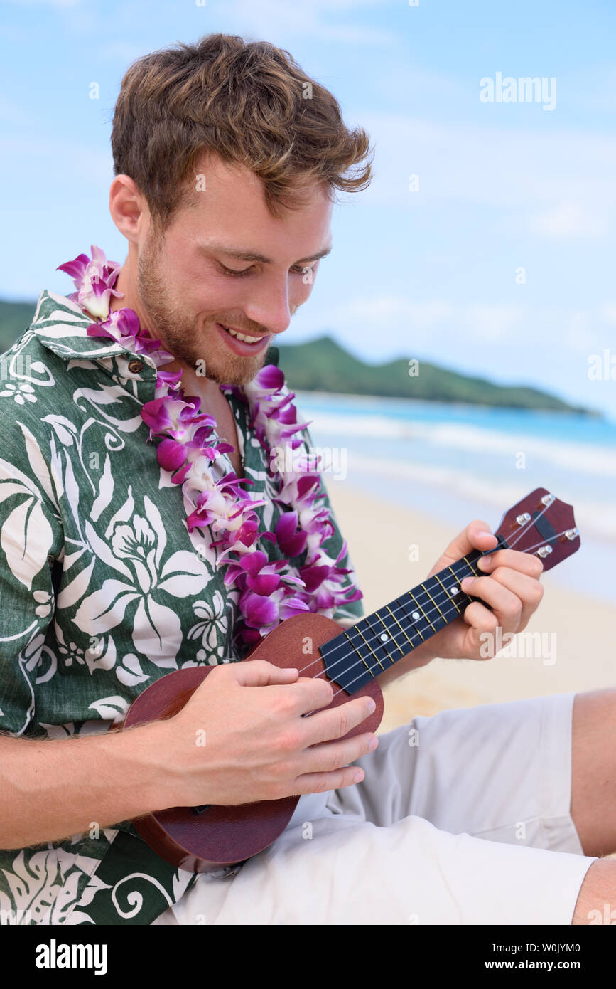 Man on beach jouer ukulele instrument sur Hawaii. Jeune homme pratiquant sur les vacances à la plage dans la région de Hawaiian portant des vêtements robe chemise Aloha et lei de fleurs. Banque D'Images