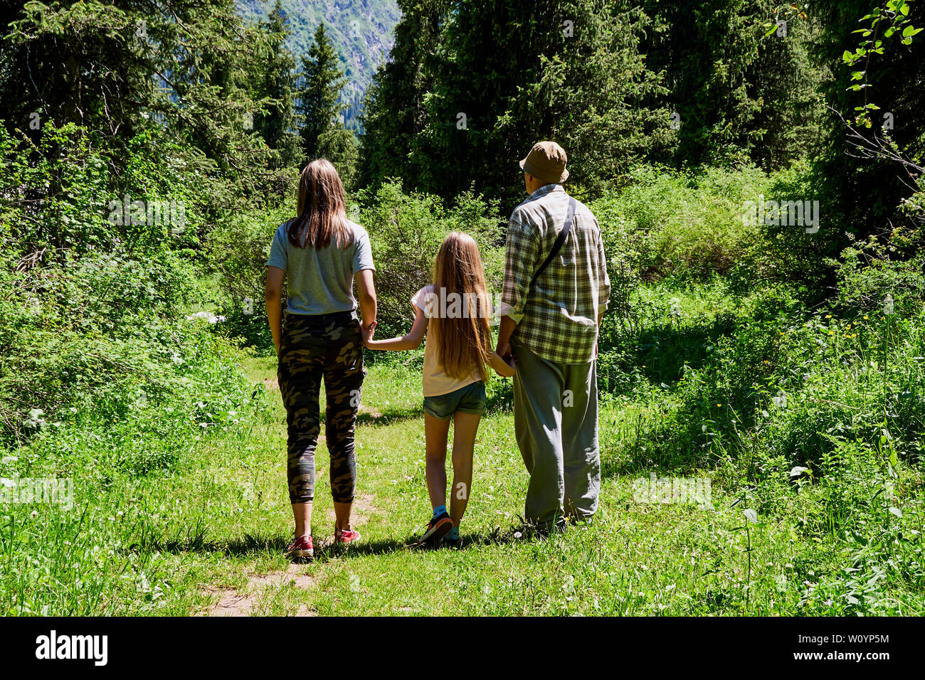 Homme adulte avec deux enfants, grand-père marche dans une forêt de montagne avec des petites-filles, journée ensoleillée d'été, fond vert, reste la randonnée Banque D'Images