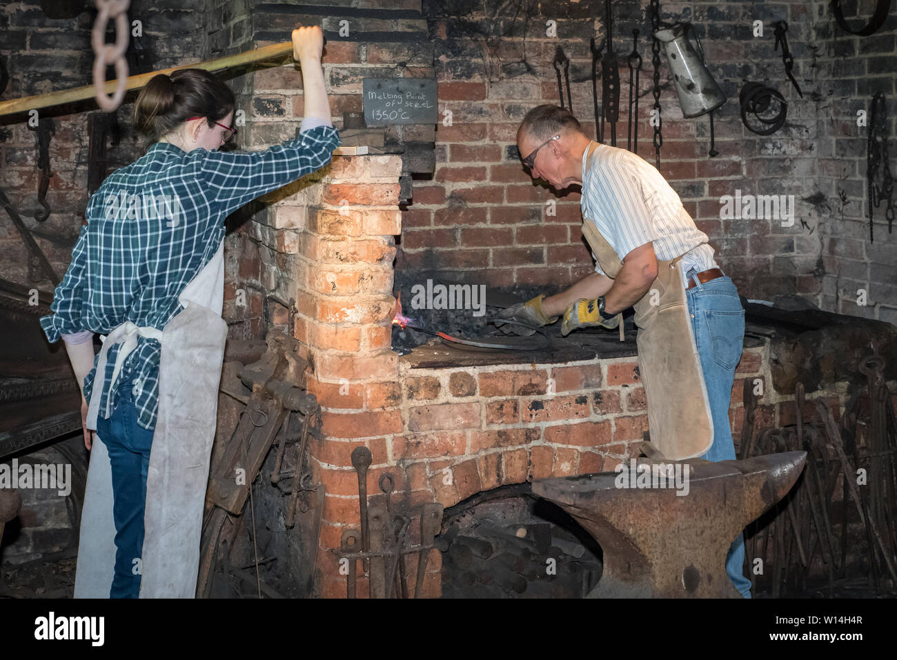 Un forgeron de sexe masculin travaillant dans une forge avec une femme pompe le soufflet. Banque D'Images
