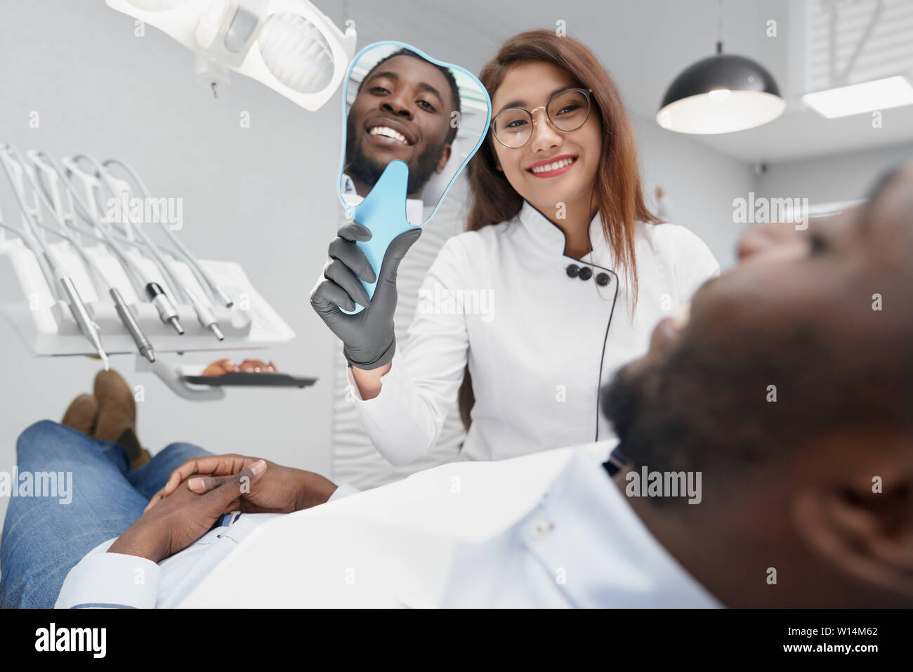 Vue du côté de happy female dentiste miroir de maintien dans la main et sourire blanc montrant à patient de sexe masculin. Jeune homme africain couché sur la chaise et dentiste Banque D'Images