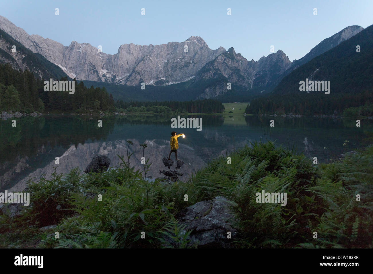 Garçon en veste jaune debout sur le rocher et tenant une lanterne à main lumineux à été lac pittoresque paysage de crépuscule, Laghi di Fusine, Italie Banque D'Images