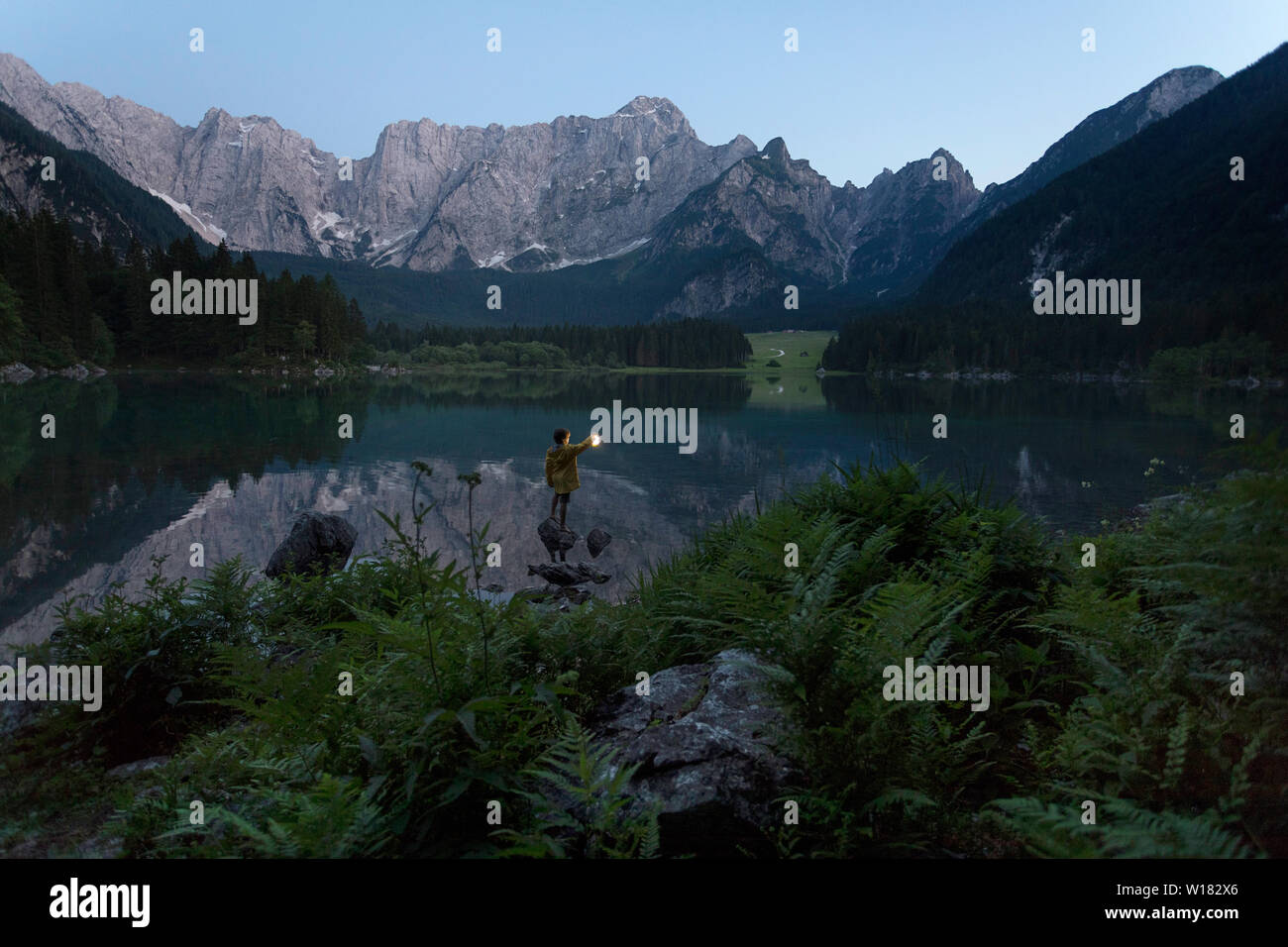 Garçon en veste jaune debout sur le rocher et tenant une lanterne à main lumineux à été lac pittoresque paysage de crépuscule, Laghi di Fusine, Italie Banque D'Images