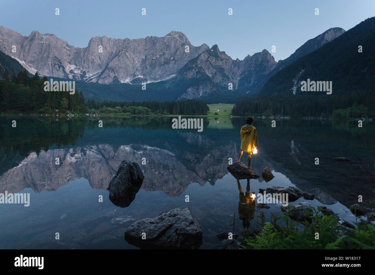 Garçon en veste jaune debout sur le rocher et tenant une lanterne à main lumineux à été lac pittoresque paysage de crépuscule, Laghi di Fusine, Italie Banque D'Images