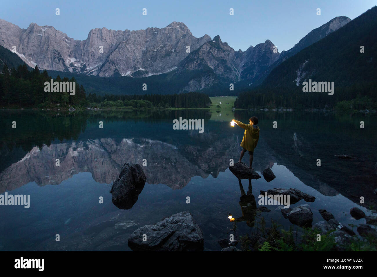Garçon en veste jaune debout sur le rocher et tenant une lanterne à main lumineux à été lac pittoresque paysage de crépuscule, Laghi di Fusine, Italie Banque D'Images