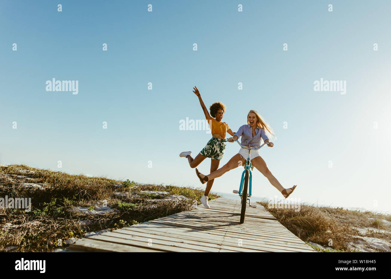 Excited woman riding bike sur la promenade avec ses amis courir derrière. Deux jeunes amis féminins ayant un grand temps sur leurs vacances. Banque D'Images