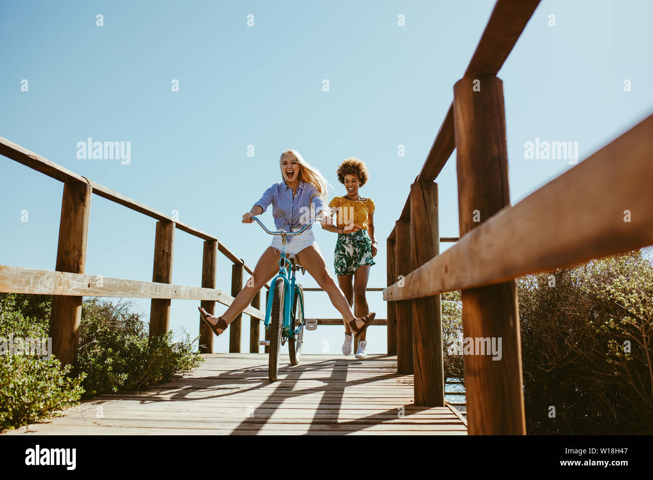 Excited woman riding bike sur la promenade avec ses amis courir derrière. Deux jeunes amis féminins ayant un grand temps sur leurs vacances. Banque D'Images
