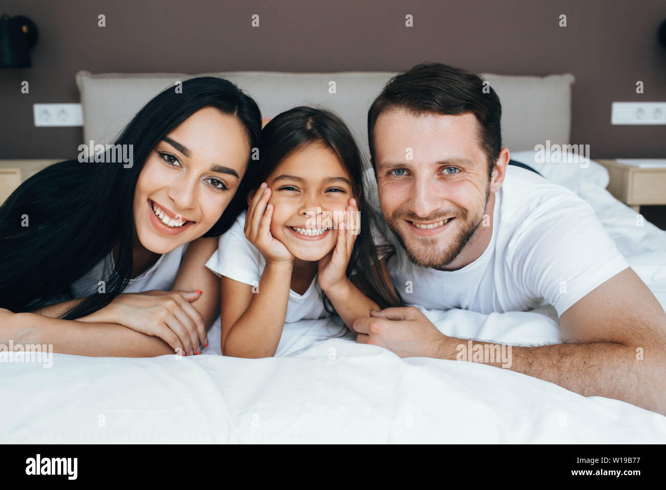 Famille heureuse allongé sur un lit blanc nouveau hugging et regardant la caméra. Mère, père et fille lying on bed at home Banque D'Images