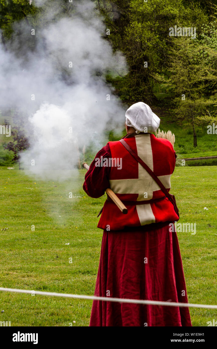 Une femme en costume médiéval anglais debout sur une pelouse au bord d'un lac et montrant une arme à feu ancienne Banque D'Images