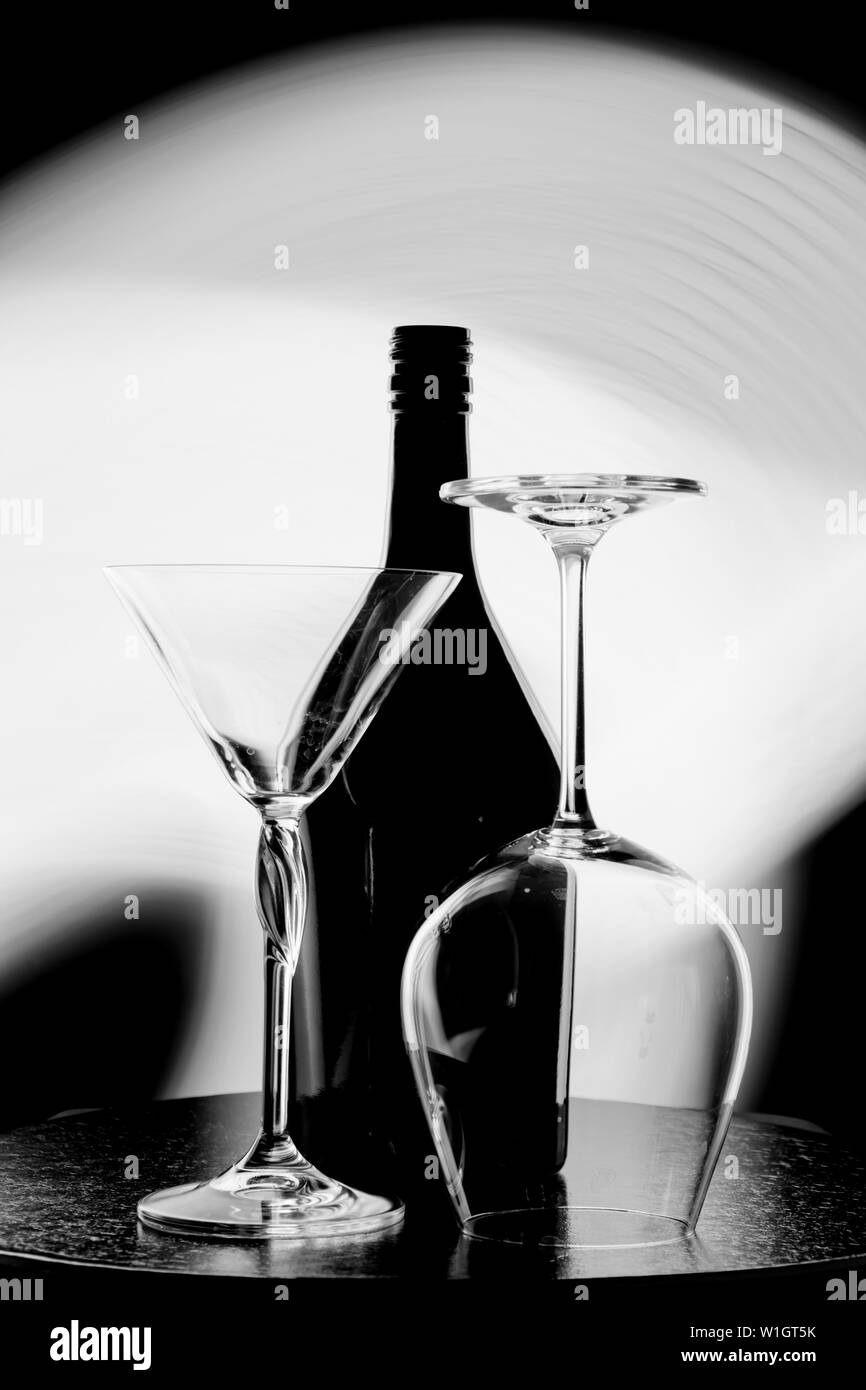 Une photo en noir et blanc d'une bouteille de vin et deux verres contre le point lumineux sur le mur noir Banque D'Images