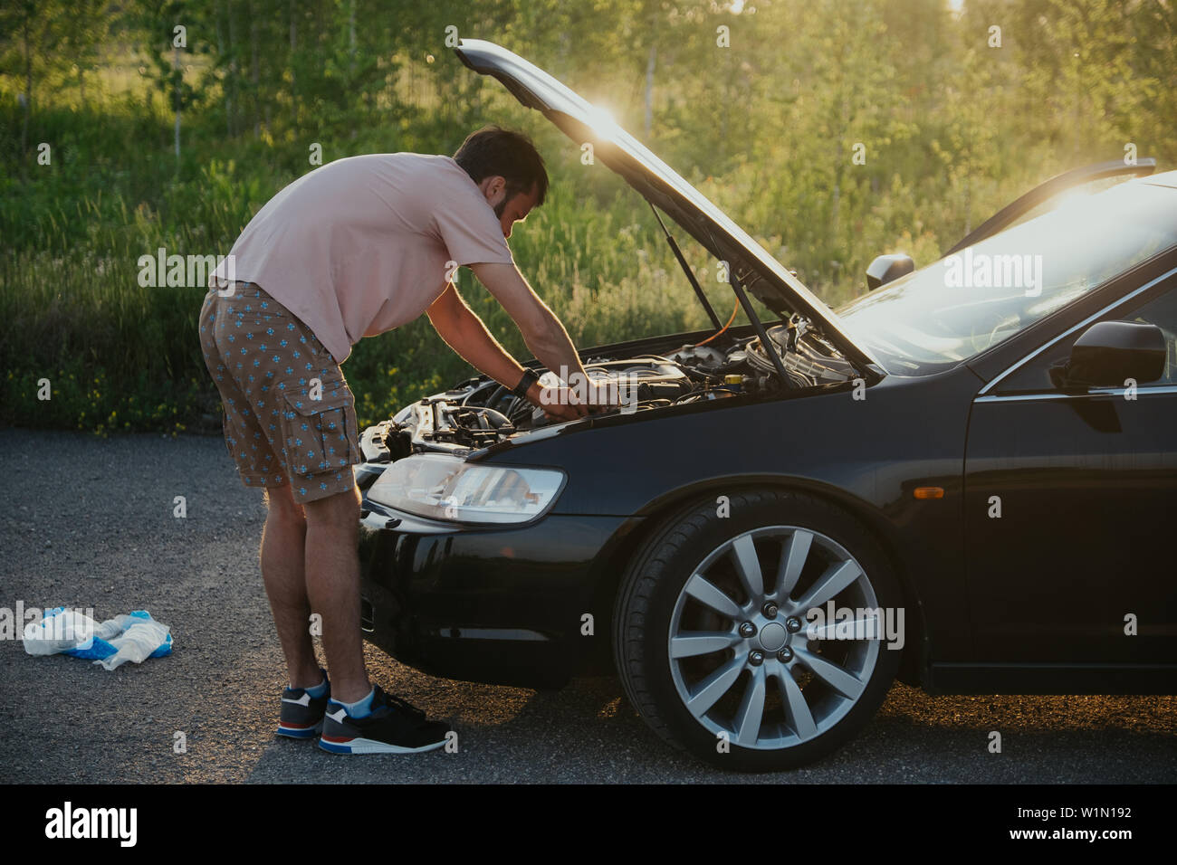 Le gars dans le t-shirt rose vérifie le moteur d'une voiture noire en soulevant le capot dans le pays à l'extérieur de la route Banque D'Images