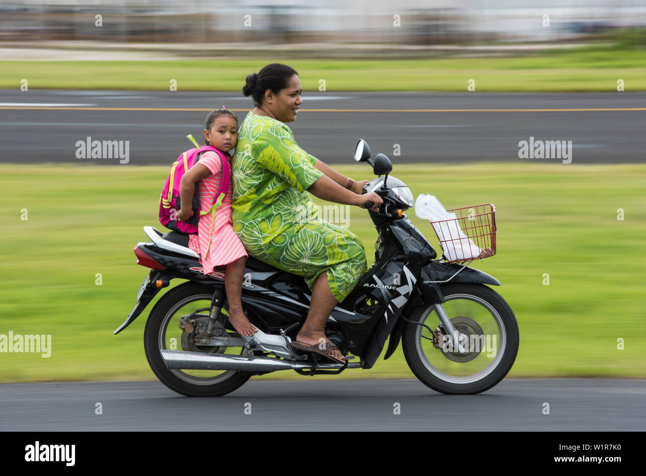 Une femme dans une robe verte conduit un motorscooter avec une fille aux pieds nus dans une robe rayée rose et blanc et un sac à dos, assis derrière elle, Fongafale Isl Banque D'Images