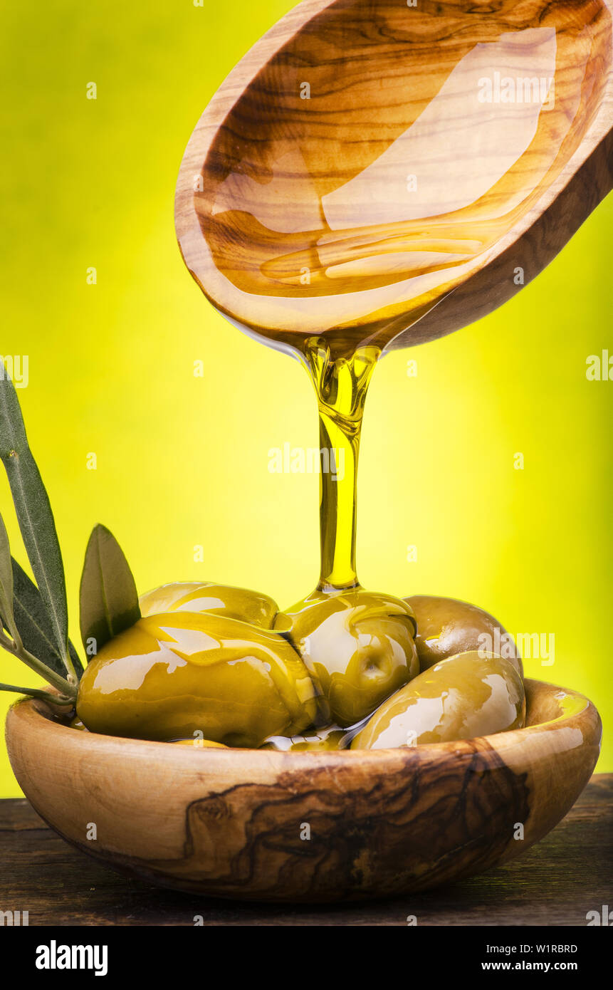 L'huile d'olive coulant dans un bol en bois d'olivier plein d'olives vertes sur fond vert. Banque D'Images