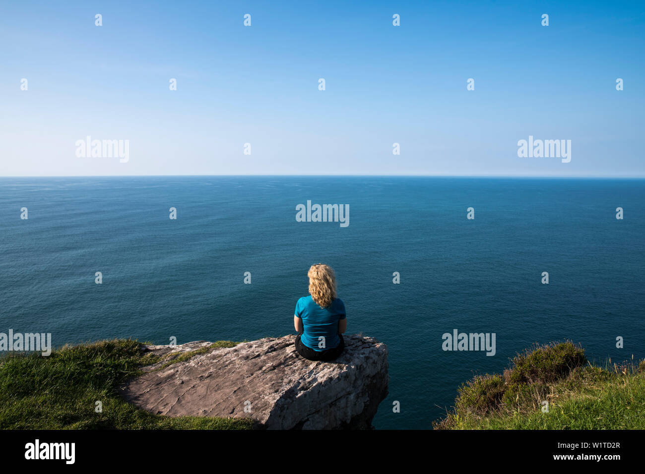 Une femme blonde s'assoit sur un rocher et surplombe la mer bleue à l'horizon, vu de tout en marchant la Dingle Way, Ballydavid Nord, Brandon, Dingle Pen Banque D'Images