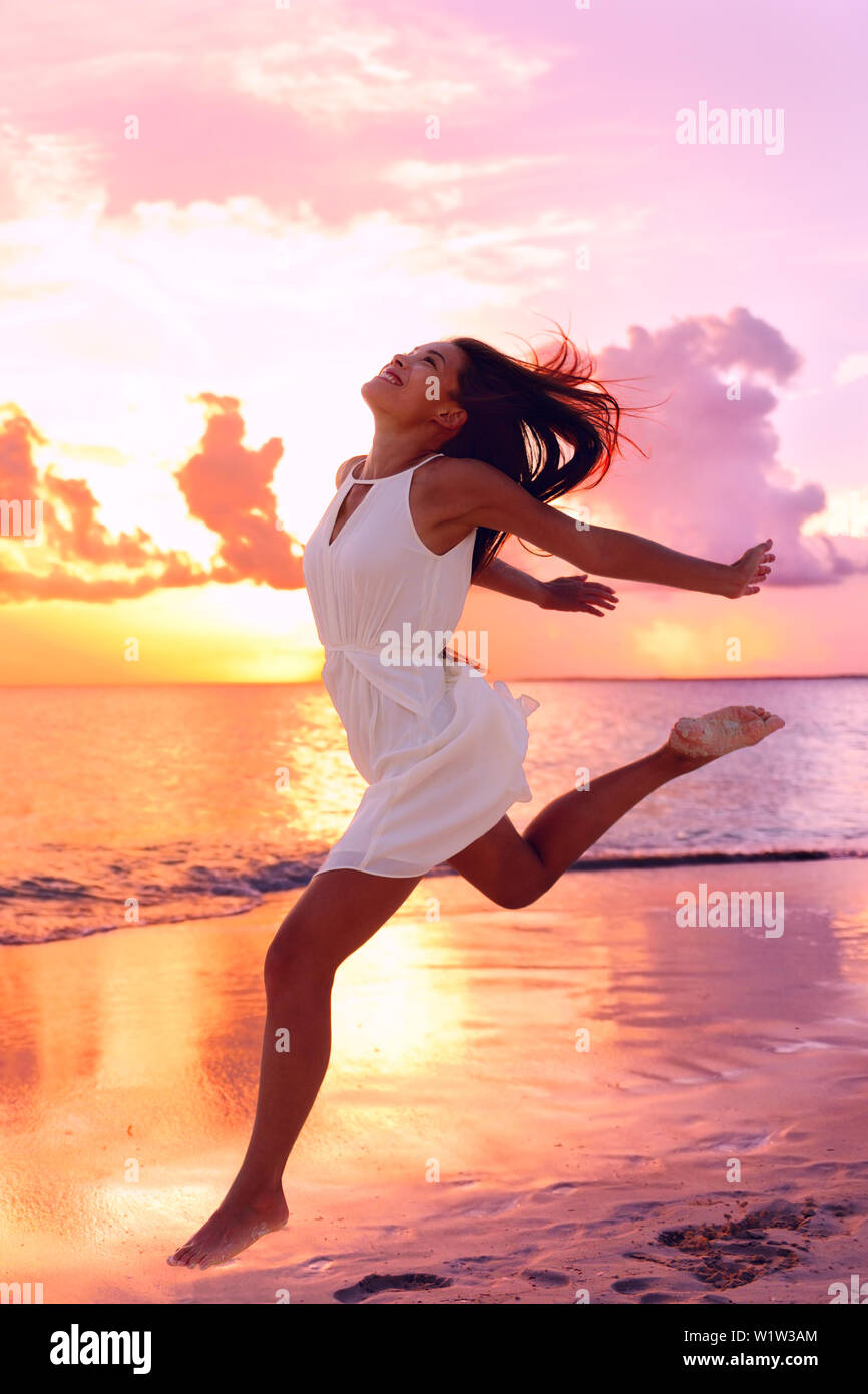 Carefree woman jumping at beach au coucher du soleil. La longueur totale de la robe d'porte. Touriste, c'est profiter de locations contre ciel orange au bord de mer. Banque D'Images