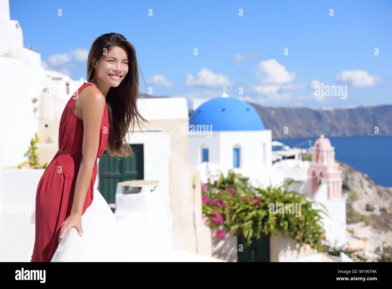 L'île de Thira Santorini Grèce tourisme - Asian woman wearing red dress on été voyage looking at view avec le célèbre attraction trois dômes chapelle église dans l'arrière-plan. Destination de luxe. Banque D'Images