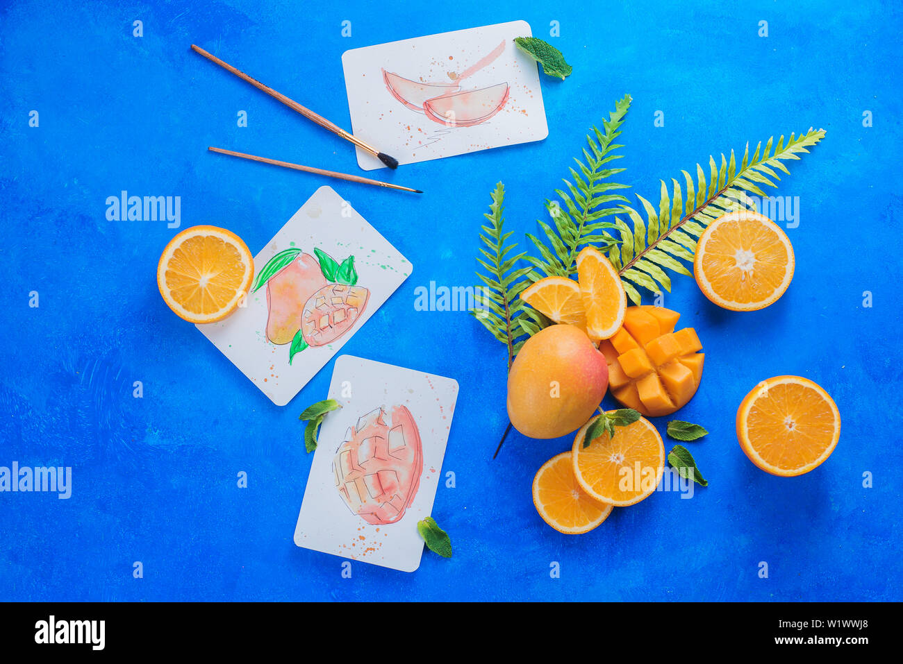Les mangues jaunes et oranges avec des feuilles tropicales et croquis à l'aquarelle sur fond bleu vif avec copie espace. Couleurs complémentaires art photographe Banque D'Images