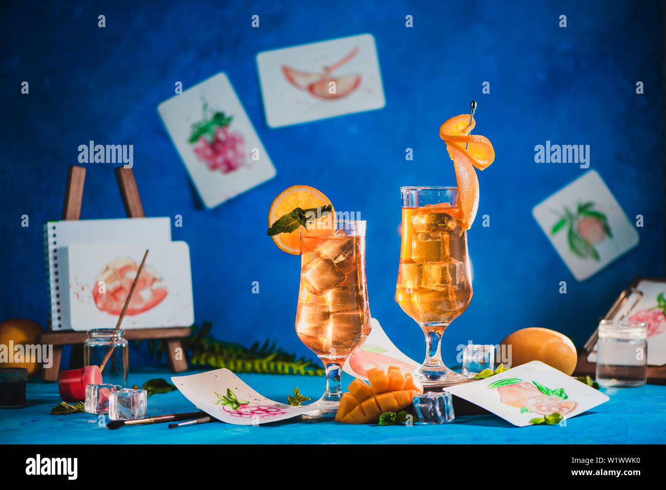 Des boissons d'été dans des grands verres avec des croquis dans un milieu de travail de l'artiste. mangue et thé glacé orange sur un fond bleu avec copie espace Banque D'Images