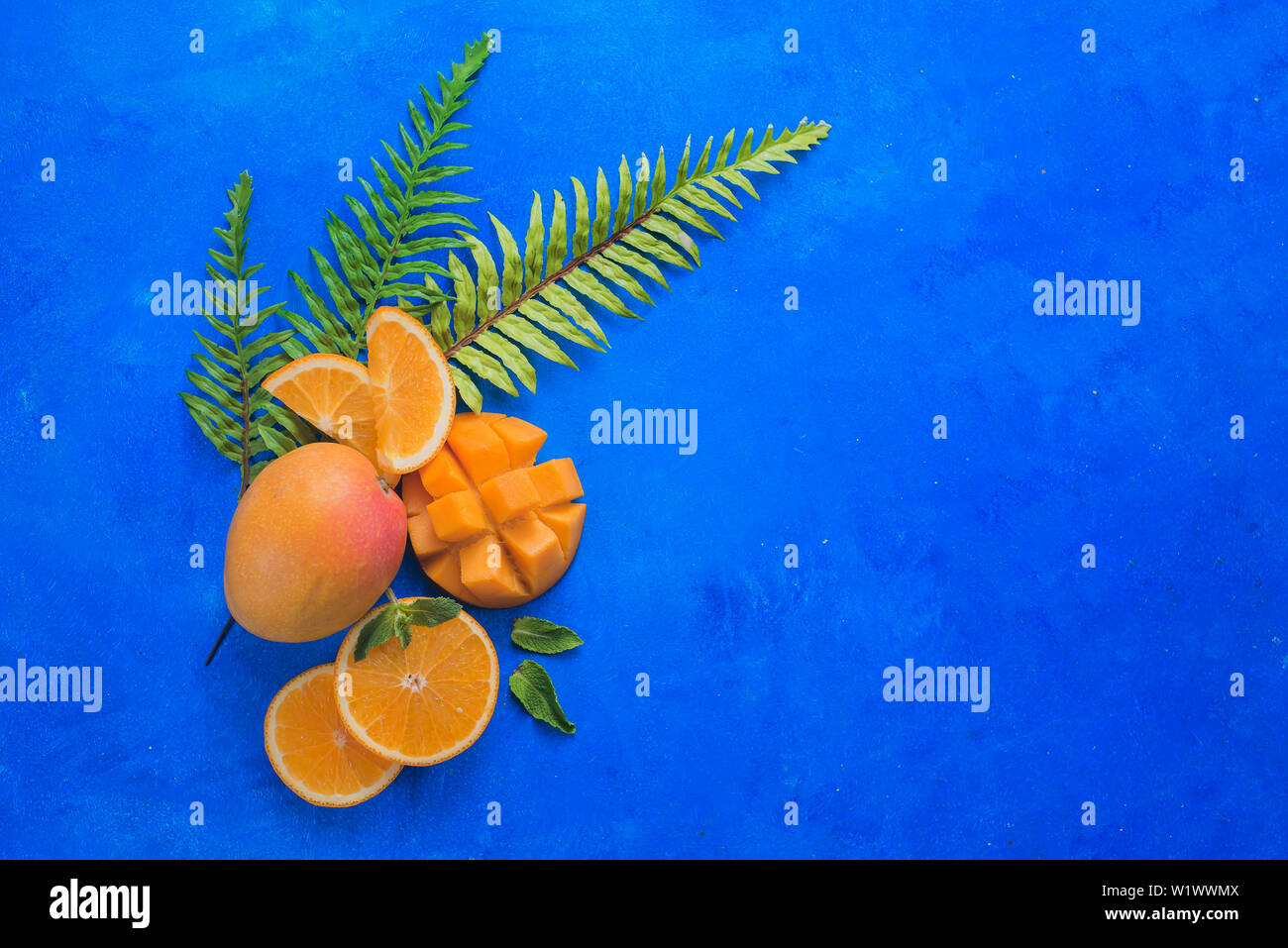 Fruits exotiques sur fond bleu vif avec copie espace. Mangue et orange avec les feuilles tropicales mise à plat. Banque D'Images
