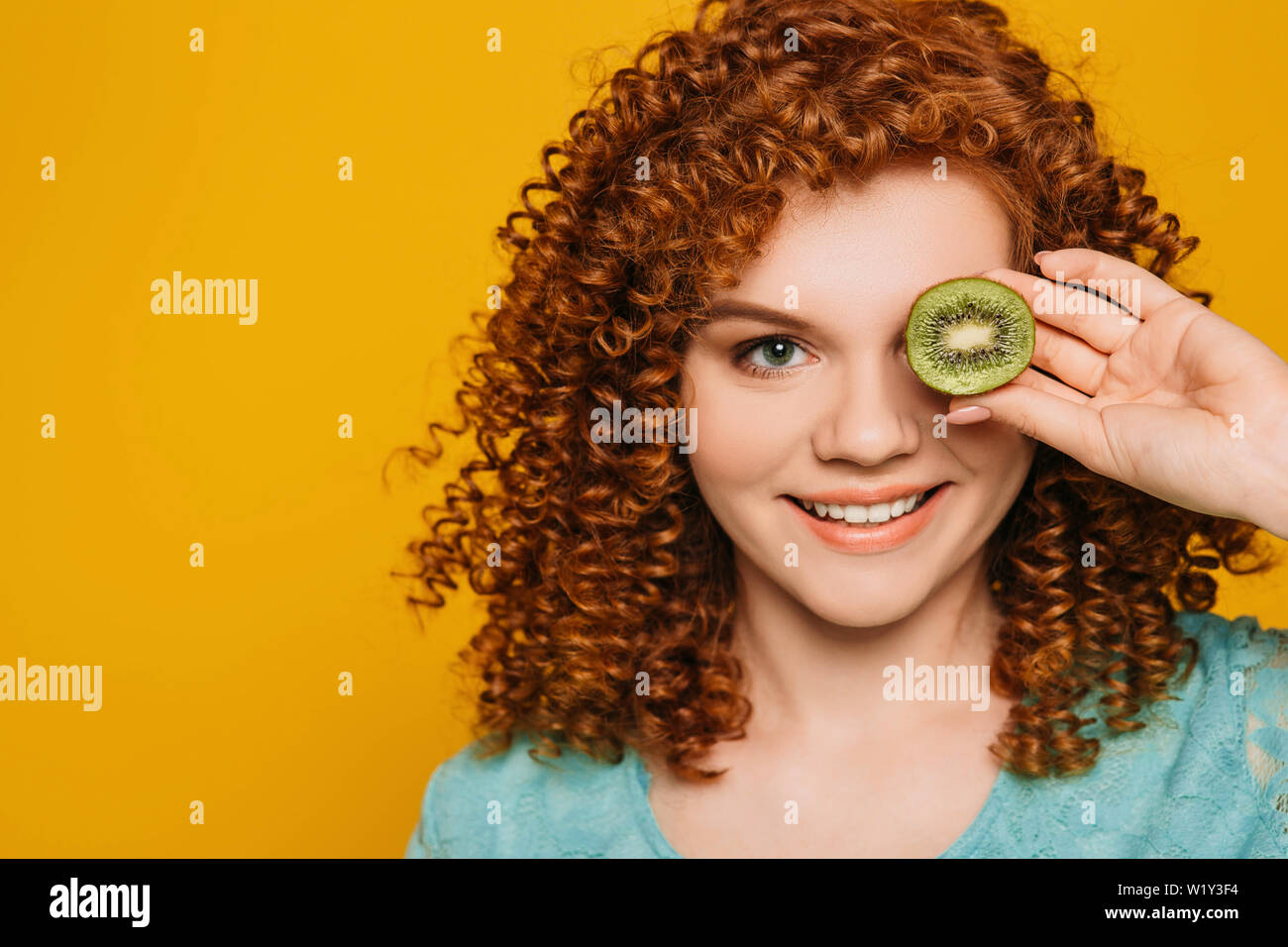 C'est un fruits detox du temps. Femme aux cheveux rouge positif avec l'oeil vert holding tranche de kiwi frais près de son œil sur mur jaune Banque D'Images