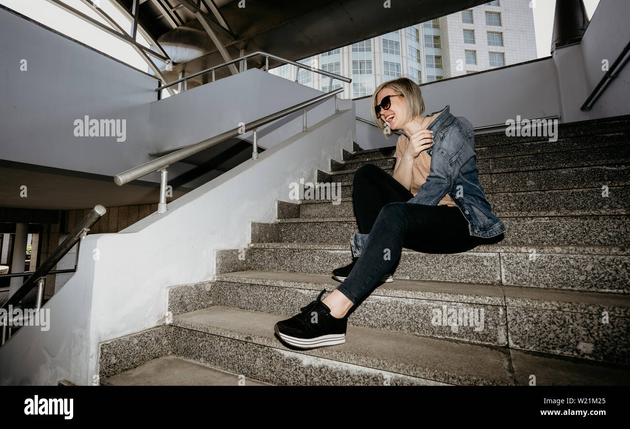Belle blonde rit assise sur les marches. Femme moderne dans la grande ville. Banque D'Images